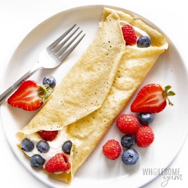 Keto crepes rolled up on a plate with whipped cream and berries.